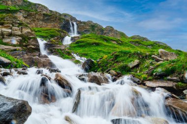 Dağlarda sersemletici bir şelale ve kayaların üzerinden akan su. Bulanık arka plan parlak yeşil tepeler ve açık mavi bir gökyüzü içeriyor. Etkileyici bir doğal manzara yaratıyor..