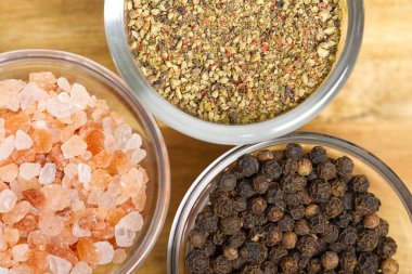 The image shows three glass bowls filled with different spices on a wooden table. One bowl is filled with pink salt, another with ground black pepper. The third bowl is filled with peppercorns. clipart