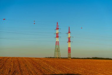 Two tall electricity pylons rise in a tranquil rural landscape at sunset. The vibrant sky colors create a stunning backdrop, highlighting the pylons against the peaceful scenery. clipart