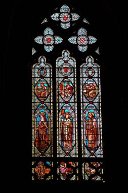 Gotik tarzda, vitraylı pencerede, bir adamın yazdığı üç bölüm, dua eden iki kadın ve karmaşık taş oymalarla çerçevelenmiş bir ayının saldırısına uğrayan bir adam var..