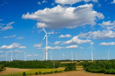 Birkaç büyük rüzgar türbininden oluşan manzaralı bir rüzgâr çiftliği temiz bir kırsal alanda temiz enerji üretir. Açık mavi gökyüzü ve tüylü beyaz bulutlar dingin bir zemin oluşturur..