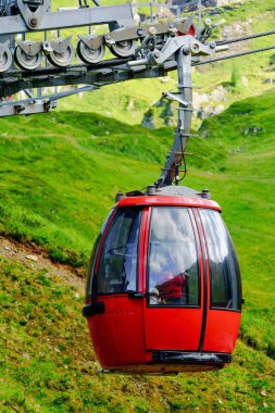 Pürüzsüz ve modern kırmızı bir teleferik yemyeşil bir dağ yamacından aşağı iniyor. Bir cam kabarcık kabini sarar ve büyüleyici manzaranın panoramik görüntüsünü sunar..