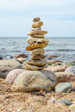 Bu resim, sakin bir plaj zeminine karşı dikkatlice dengelenmiş pürüzsüz, yuvarlak taşlar sergiliyor. Sakin mavi deniz yavaşça kıyıda döner, gökyüzü berrak ve davetkar kalır..