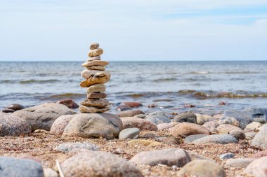 Sahilde arka planda deniz olan dengeli taşlar yığını. Taşlar pürüzsüz ve yuvarlaktır ve su koyu mavi renktedir. Gökyüzü açık mavi ve beyaz bulutlar.