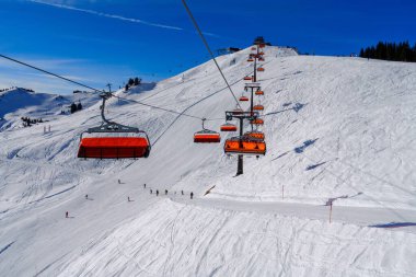 Canlı kırmızı ve beyaz bir kayak asansörü Avusturya 'nın pitoresk Alplerinde karla kaplı bir dağa tırmanır. Parlak güneş ışığı ve açık gökyüzü çarpıcı bir kış sporu atmosferi yaratır..