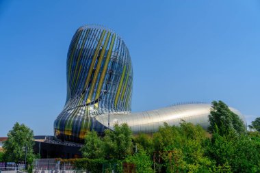 Canlı yeşil ağaçlarla çevrili, yansıtıcı panelleri ve çarpıcı mimarisi olan modern bir metalik müze, parlak mavi gökyüzüne karşı kurulmuş, çarpıcı bir görsel deneyim yaratıyor..