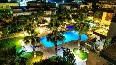 Tropikal bir gecenin cazibesini yaşayın. Palmiye ağaçlarıyla çevrili güzel aydınlatılmış bir tatil köyü havuzu. Bu resim, yıldızlı bir gökyüzünün altında huzurlu bir tatil kaçamağının özünü yakalar..