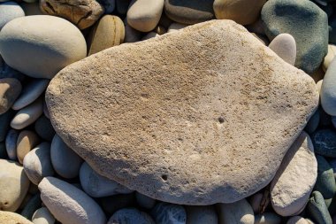 Bu resimde, sakin bir sahilde pürüzsüz çakıl taşlarıyla çevrili büyük, dokulu bir taş görülüyor. Güneş ışığı kıyı güzelliğini artırarak, doğa temalı tasarımlar için ideal hale getiriyor..