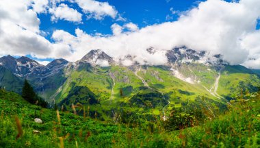 Pofuduk beyaz bulutları olan parlak mavi gökyüzünün altındaki yeşil dağların çarpıcı manzarası. Bu resim doğanın dingin güzelliğini yakalar, seyahat ve manzara için idealdir..