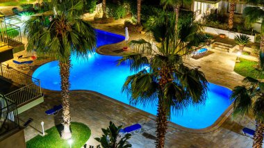Palmiye ağaçları ve rahat şezlonglarla çevrili güzel mavi yüzme havuzlu, tatil promosyonları için ideal, sakin bir gece manzarasının tadını çıkarın..