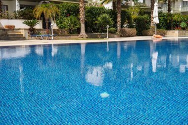 Çevredeki binaları ve yeşillikleri yansıtan berrak mavi yüzme havuzunun çarpıcı manzarası. Sahne rahatlama ve sükuneti çağrıştırıyor. Yaz promosyonları ve tatil kavramları için mükemmel..