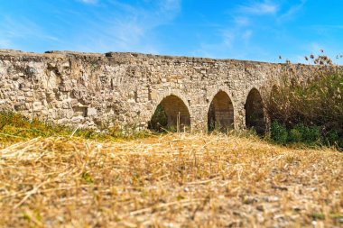 Ele geçirilen, temiz mavi gökyüzüne karşı dizilmiş üç kemerli eski bir taş su kemerinin resimli görüntüsü. Kuru çimen manzarası tarihsel cazibesine ve ebedi güzelliğine katkıda bulunur..