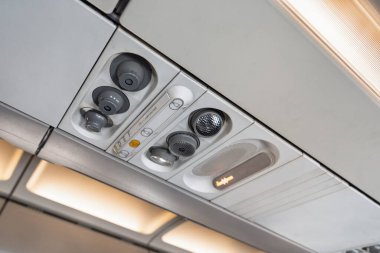 Yukarıda ayarlanabilir havalandırma delikleri, okuma ışıkları ve bir çağrı düğmesi gösteren bir uçak konsolu var. Bu resim hava yolculuğu rahatlığını ve yolcu kolaylığını vurgular.