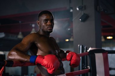 Siyah boks eldivenli kaslı boksör ringde kolları ve kum torbasıyla duruyor..