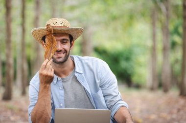 Hasır şapkalı, dizüstü bilgisayarlı ve doğadaki kameraya bakan gülümseyen bir adamın portresi..