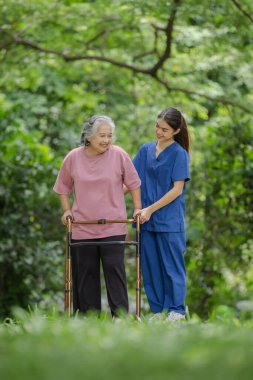 Yürüyen yaşlı bir kadın yemyeşil bir park ortamında genç bir hemşire tarafından dikkatlice destekleniyor.