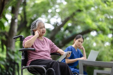 Elderly woman in a wheelchair expressing concern during a conversation, while a nurse looks on, in a serene outdoor setting. clipart