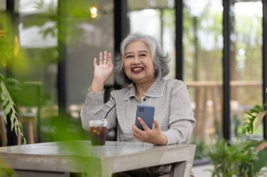 Cafe 'de Mutlu Yaşlı Kadın El sallıyor ve Akıllı Telefon Kullanıyor: Kahve Bağlanıyor, Eğleniyor ve Teknolojiyi Kucaklıyor