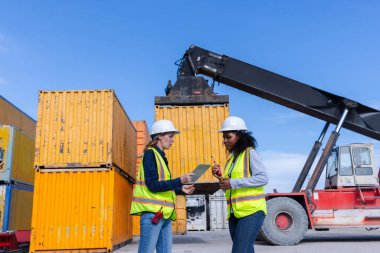 Two Female Engineers in Safety Vests Oversee a Shipping Yard Operation with a Container Lift Handling Cargo, Ensuring Safety and Efficiency in Freight Management and Logistics Operations clipart