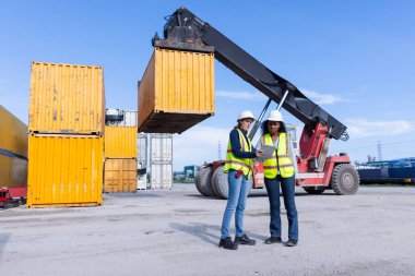 Two Female Engineers in Safety Vests Oversee a Shipping Yard Operation with a Container Lift Handling Cargo, Ensuring Safety and Efficiency in Freight Management and Logistics Operations clipart