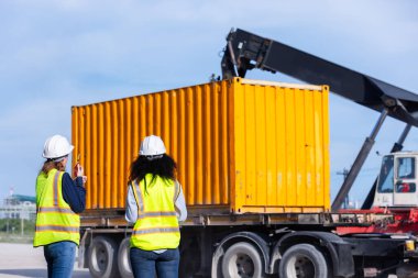 Two Female Engineers Supervising the Loading of a Yellow Shipping Container onto a Truck Using Heavy Machinery at a Shipping Yard, Ensuring Safe and Efficient Logistics Management clipart