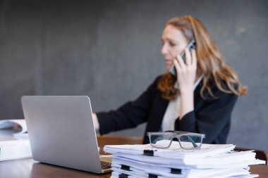 Focused Businesswoman Reviewing Reports and Handling a Phone Call in a Professional Office Setting clipart