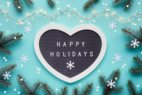 テキストハッピーホリデーフェルトハート形のレターボード レターボード上 冬のクリスマスは モミの小枝 紙の雪のフレーク クリスマスライト付きのガーランドで背景を平らにします モノクロマティックブルーミントの背景 — ストック写真