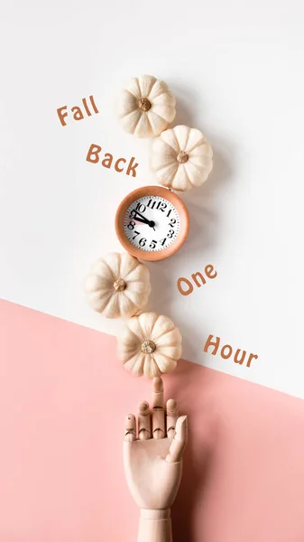 Time change in Autumn. Text Fall Back 1 hour on circle stone board. Wooden alarm clock, with dry Fall leaves on recycled cardboard. Top view, monochromatic beige, pink flat lay, soft natural sunlight.