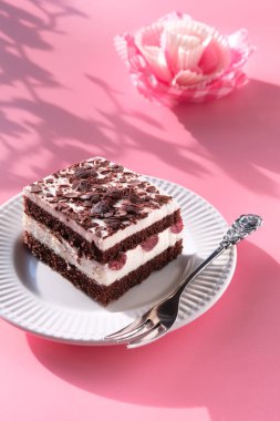 Kirazlı çikolatalı pasta. Çatallı bir tabakta çocuk oyuncağı. Canlı pembe arka planda, kağıt çiçekli, el yapımı çiçek desenli tatlı. Gölgeli güneş ışığı.