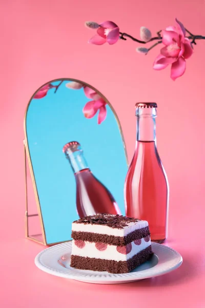 Chocolate cake with cherries. Piece of cake on a plate with fork. Bottle of pink vine reflected in arch mirror. Sweet dessert on pink background with plum blossoms. Slice of torte and Spring flowers .