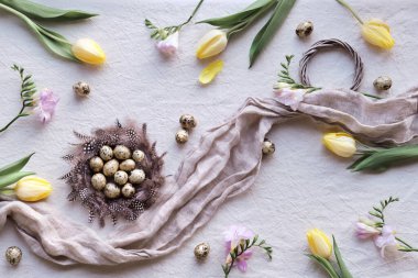 Natural Easter decor, overhead, flat lay with quail eggs in feather nest, yellow tulips and pink freesia flowers on off white textile background. Flat lay, top view springtime background..