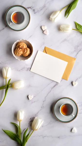 Blank postcard, envelope, white tulips and tea cups with heart-shaped sugar lumps on a marble background. Top view, copy space, place for text. Perfect spring background for social media stories