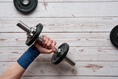 Teenage hand, dumbbel on wooden floor, flat lay, top view. Wrist wrapped in blue elastic bandage.