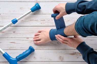 Teenage boy wrapping compression bandage around sprained ankle to reduce ache. Crutches on the floor. Feet trauma, injury. Medical Ankle Bandage. Ankle Medical Support Strap, Adjustable Wrap Bandage.