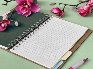 Yanında bir kalem olan bir defterin fotoğrafı. Yanında da canlı bir çiçek dizilimi. Hepsi bir masanın üzerinde duruyor. Pembe manolya çiçekleri.