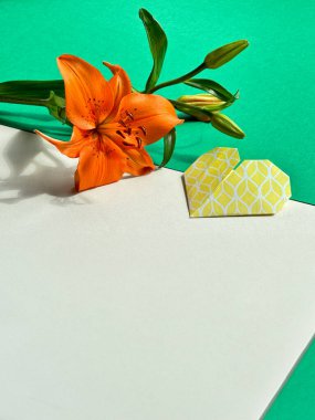 Canlı bir turuncu zambak, zıt yeşil ve beyaz arka planda sarı origami bir kalp ile eşleştirildi. Kart tasarımları, davetiyeler ve dekoratif posterler için mükemmel..