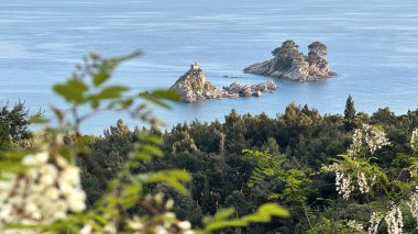 Karadağ 'da yemyeşil ve çiçek açan bitkilerle çerçevelenmiş sakin mavi sularla çevrili küçük kayalık adaların yer aldığı resim gibi bir kıyı manzarası. Petrovac, Katic Adaları ve Sveta Nedjelja