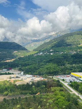Karadağ 'ın Tivat kenti yakınlarındaki yeşil arazi, dağınık binalar ve görkemli dağları gösteren hava perspektifi. Sahnede parçalı bulutlu bir gökyüzü altında doğal yeşillik ve kentsel gelişimin bir karışımı yer alıyor..