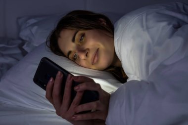 Karanlık bir odada, beyaz bir yatakta telefonu olan bir kız.
