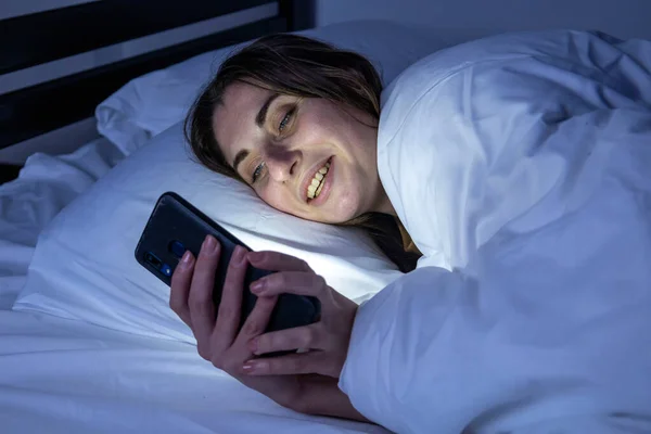 A girl in bed with a phone on a white bed in a dark room