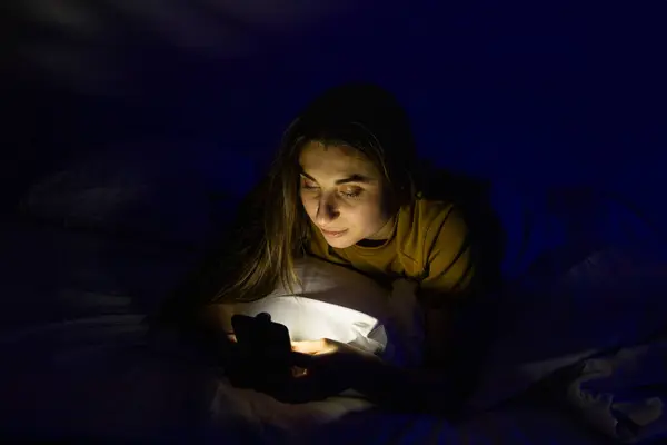 stock image A girl in bed with a phone on a white bed in a dark room