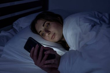 Karanlık bir odada, beyaz bir yatakta telefonu olan bir kız.