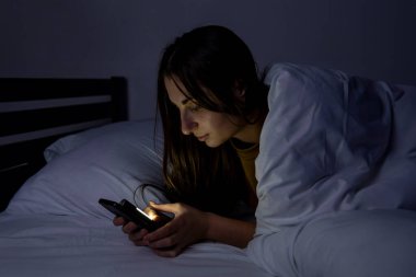 Karanlık bir odada, beyaz bir yatakta telefonu olan bir kız.