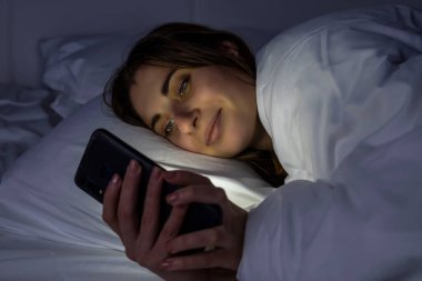 Karanlık bir odada, beyaz bir yatakta telefonu olan bir kız.