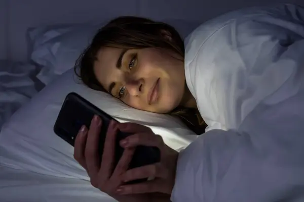 A girl in bed with a phone on a white bed in a dark room