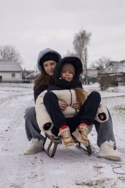 İki kız kardeş kış eğlencesinin tadını çıkarıyorlar, karlı bir arazide tahta bir kızağın üzerinde oturuyorlar, sıcak kışlık giysiler giyiyorlar.
