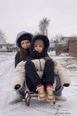 İki kız kardeş, karlı bir kırsalda tahta bir kızağın üzerinde otururken fotoğraf için poz veriyorlar.