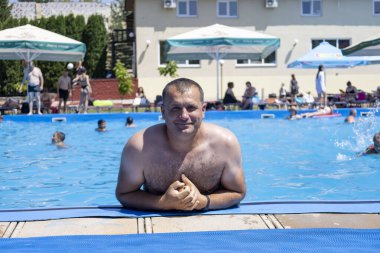 Sıcak bir yaz gününde havuzun kenarında dinlenen gömleksiz bir adam ferahlatıcı suyun ve diğer yüzücülerin arkadaşlığının tadını çıkarıyor.