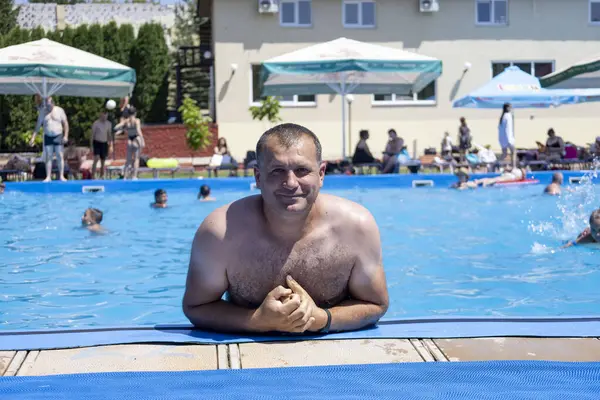 Sıcak bir yaz gününde havuzun kenarında dinlenen gömleksiz bir adam ferahlatıcı suyun ve diğer yüzücülerin arkadaşlığının tadını çıkarıyor.