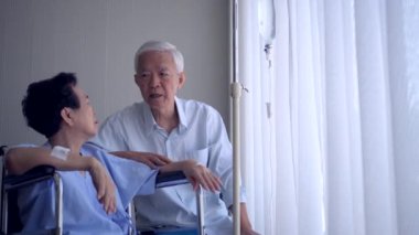 Asyalı yaşlı çift tekerlekli sandalye yaşı ve sağlık promblemi üzerine hastanede konuşan eşi destekliyor.